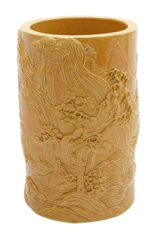 An ochre glazed porcelain brush pot, of cylindrical form, carved in relief with seated buddha in a river landscape, four character Qianlong seal mark, 12.5cm high.