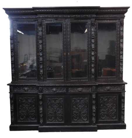 A Victorian carved oak breakfront bookcase, with an out swept pediment, over four glazed doors, flanked by lion's head and trailing fruit, enclosing two long and four short shelves, above a base section with four mask and foliate carved handled frieze dra