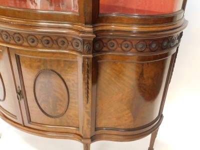 An early 20thC mahogany display cabinet or vitrine, the top with a shaped edge and a frieze carved with oval and circular patera above a serpentine shaped glazed door flanked by shaped sides, enclosing a silk lined interior, the base with two panelled doo - 2