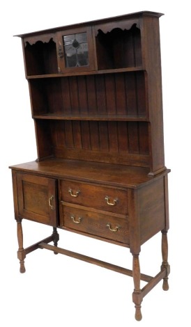 A late Victorian oak dresser, the outswept pediment over a stained glass central door, flanked by recesses, on a two shelf plate rack, above a cupboard door flanked to the right by two frieze drawers, raised on turned legs united by a h frame stretcher, 1