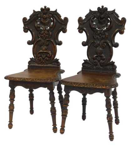 A pair of Continental carved oak hall chairs, each with a scroll leaf and floral carved back, solid seat with carved border, on leaf carved turned cylindrical legs.