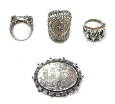 A silver Lincoln Cathedral bar brooch, 4cm wide, and three Eastern inspired dress rings, one formed and four wolves, the other florally decorated, white metal unmarked, 23.3g all in. (4)