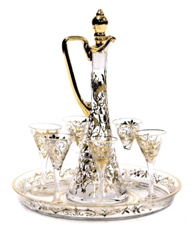 An early 20thC cut glass liqueur set, decorated in gilt and white metal overlay, with trailing flowers and leaves, comprising a liqueur jug and stopper, six glasses and a circular tray.