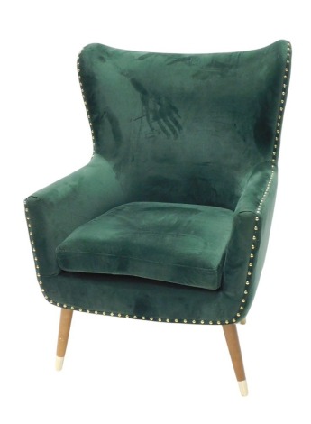 A green velvet upholstered wing back chair, with brass studded borders, and turned legs.