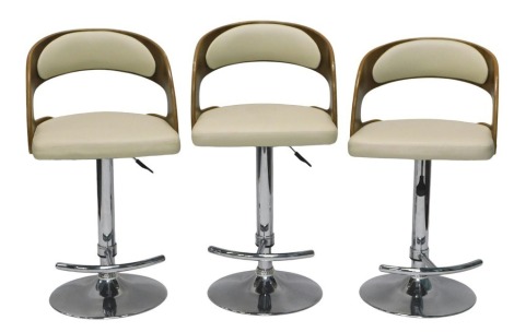 A set of three bar stools, each with a walnut veneered back, a cream leatherette padded seat and back rest, on a chrome base with foot rest.