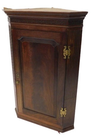 A George III mahogany hanging corner cabinet, with a blind fret frieze above a panelled door with elaborate pierced H shaped hinges, flanked by reeded canted sides, 118cm high, 79cm wide.