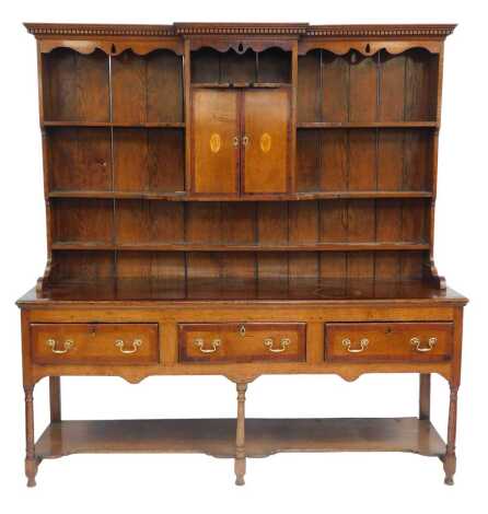 A 19thC oak dresser, the top with a dentil moulded cornice, above a recess with two cupboard doors beneath inlaid with shell paterae flanked by two shaped shelves and an under shelf, the base with three drawers above a shaped under tier, raised on turned 