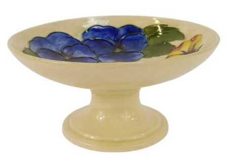 A Moorcroft pottery pedestal dish decorated in the Pansy pattern, against a pale yellow ground, impressed marks and partial paper label, 8.5cm high, 18cm diameter.