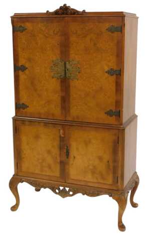 A Queen Anne style walnut cocktail cabinet, the burr wood doors with cast metal ornate floral cast metal hinged and lockplate, enclosing a mirrored back, two glass shelves above sliding drawer with shelves to each door, the base with two cupboard doors en