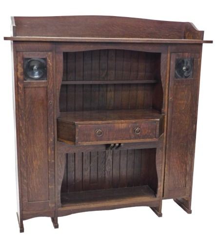 A 1920's Liberty & Co Arts and Crafts oak cabinet, with gallery top, open centre section below with a narrow shelf and break front drawer with two iron ring handles, flanked by slender cupboards, with panelled doors and bulls eye glass inserts, the open c