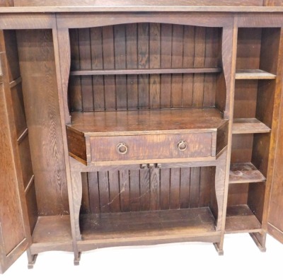 A 1920's Liberty & Co Arts and Crafts oak cabinet, with gallery top, open centre section below with a narrow shelf and break front drawer with two iron ring handles, flanked by slender cupboards, with panelled doors and bulls eye glass inserts, the open c - 2