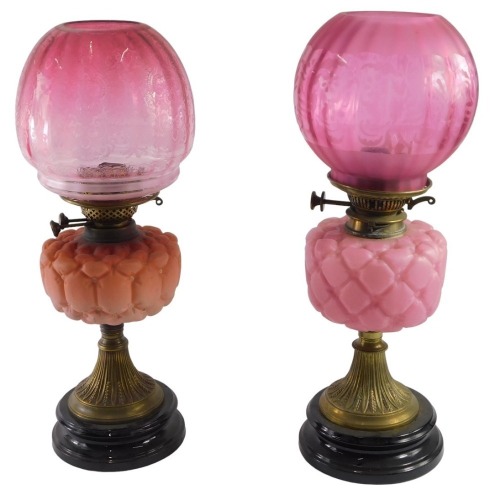 A pair of late Victorian brass oil lamps, with coloured satin glass button cushion effect reservoirs, one in pink and one in orange, with twin burners, and both on black ceramic socles, each with a cranberry glass shade having acid etched decoration.