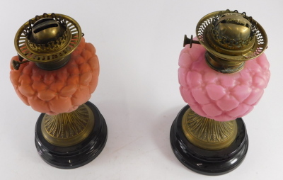 A pair of late Victorian brass oil lamps, with coloured satin glass button cushion effect reservoirs, one in pink and one in orange, with twin burners, and both on black ceramic socles, each with a cranberry glass shade having acid etched decoration. - 2