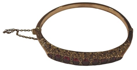 A Victorian 9ct gold ruby and diamond bangle, of half hinged design, the raised top set with nine oval rubies, with tiny diamond breaks, in scroll setting, yellow metal stamped 375, Birmingham 1883, of hinge design with safety chain, 6.5cm diameter, 10g.