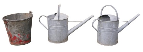 Three galvanised wares, comprising two watering cans and a fire bucket.