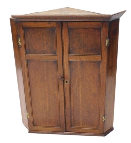 A George III oak hanging corner cupboard, dental cornice, canted sides, with three shaped shelves, enclosed by two panelled doors, with brass H hinges, 100cm high, 85cm wide. (AF)