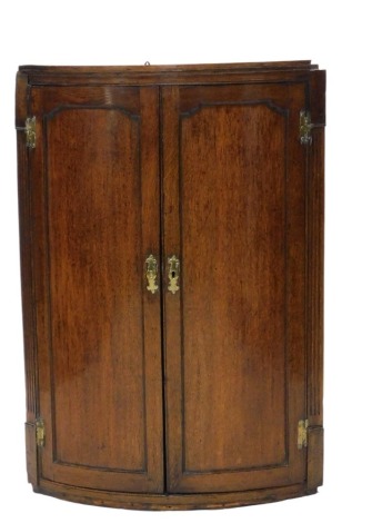 A Georgian oak cylinder fronted hanging corner cupboard, with plain panelled doors and reeded flat pilasters, brass lock plate and hinges, and enclosing three shelves within, 102cm high, 72cm wide.