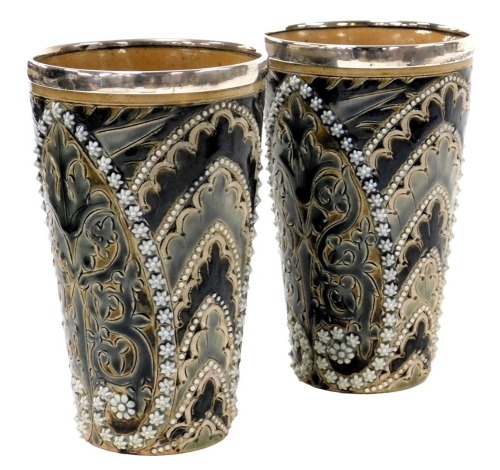 A pair of late 19thC Doulton Lambeth stoneware beakers, with silver mounts, sgraffito decorated with stylised leaves, and moulded with repeating flower heads and beading, impressed marks, London 1889, 14cm high.