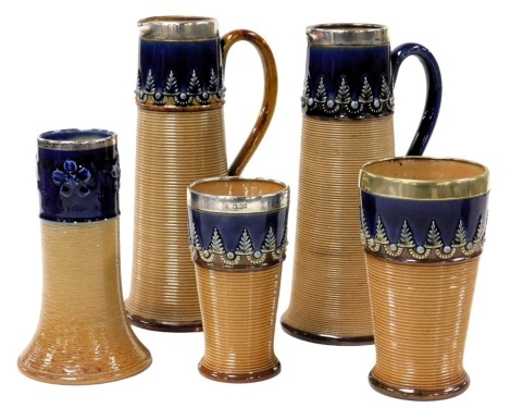 A group of late 19thC Doulton Lambeth Stoneware pottery, with silver or plated rims, relief decorated with leaves or flowers, above a ribbed lower body, impressed marks, comprising two jugs, 21cm high, two beakers and a vase.