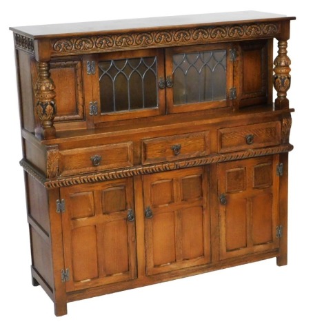 A late 20thC oak Elizabethan style court cupboard, with a carved frieze over a pair of leaded glass doors, raised on leaf carved cup and cover supports, the base with three frieze drawers over panelled doors, raised on stiles, 137cm high, 137cm wide, 46cm