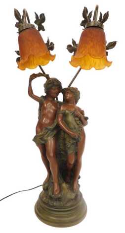 A 20thC painted cast metal figural table lamp, with two amber glass shades, the base modelled as young male and female scantily clad, on a socle base, 74cm high.