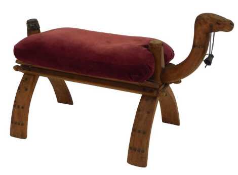 A 20thC camel stool, carved with a camel's head, with reins and bell, with a loose red draylon upholstered cover, 82cm wide.