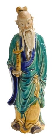 A 19th/20thC Chinese part glazed ceramic figure of a sage, in green, blue and yellow; holding a sheathed sword, 31cm high.