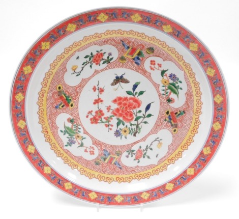 A 20thC Chinese porcelain charger, with butterfly and blossom central reserve enclosed by floral cartouches and precious objects, on a red diaper ground with yellow border, the rim having a red enamel ground with repeated floral decoration, the base signe