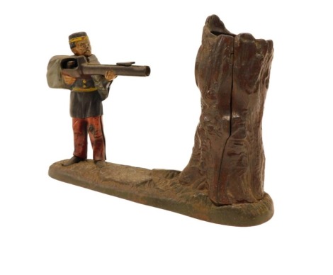 A cast iron money box, modelled in the form of a soldier shooting a rifle at a target mounted on a tree stump, indistinct patent button, bearing date 1875 to underside and indistinctly titled Creedmore Bank, 23cm wide.