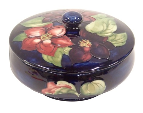 A Moorcroft ceramic squat shaped jar and cover, decorated with purple and pink flowers and leaves, on a blue ground, impressed marks to underside and paper label, 17cm diameter.