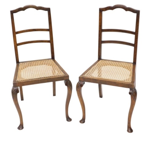 A pair of early 20thC walnut bedroom chairs, each with a ladder back and cane seat, on shell carved cabriole legs with pad feet.