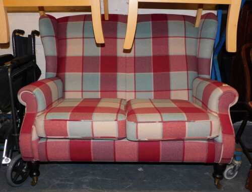 A compact two-seater wing back sofa, raised on turned hardwood legs, on brass castors, finished in a grey and red tartan fabric, overall size 125cm wide x 1000cm high x 95cm deep.