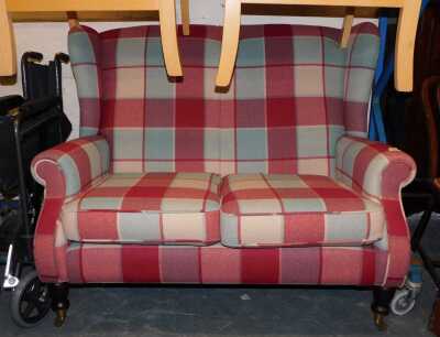 A compact two-seater wing back sofa, raised on turned hardwood legs, on brass castors, finished in a grey and red tartan fabric, overall size 125cm wide x 1000cm high x 95cm deep.