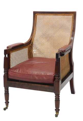 A Regency mahogany bergere library chair, with slope back and red leather padded arm rests and seat, and loose cushion, on turned tapering fluted legs and brass castors. Upon initial inspection there is a section of damage to the caning bottom left corner