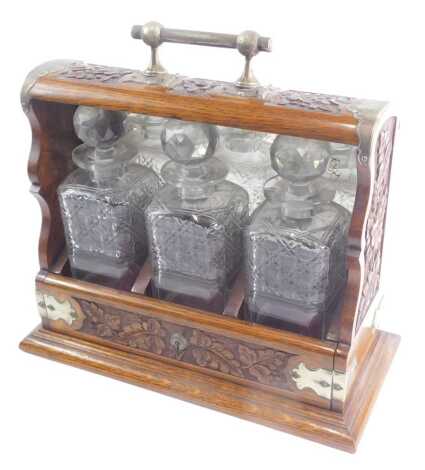 An Edwardian carved oak tantalus, with silver plated mounts, containing three square section cut glass decanters and stoppers, and with mirrored back, 37cm wide.