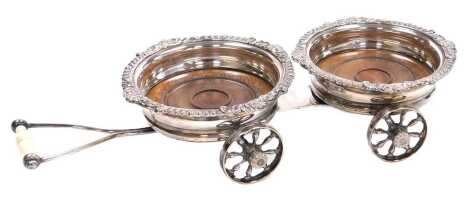 A late 19thC silver plated double bottle coaster trolley, each coaster with a gadrooned and foliate rim, with a mahogany base, the trolley with four floral capped wheels, and a turned ivory handle, 44cm long.