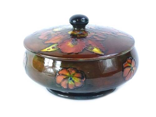 A Moorcroft Flambe Orchid pattern box and cover, on a mottled blue and brown ground, impressed mark and paper label to underside, 15cm diameter.