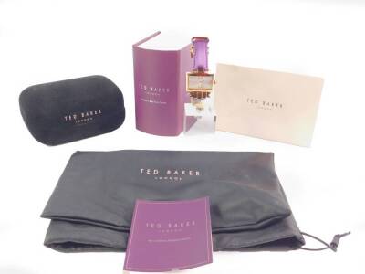 A Ted Baker lady’s gold plated dress wristwatch, rectangular silvered dial bearing Roman numerals at 12 and 6, Arabic numeral at 3, on a lilac strap, with case and booklet, together with a Ted Baker jewellery/glasses case, and a drawstring bag.