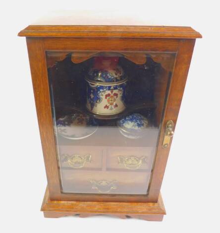 A Victorian oak smoker’s cabinet, with a bevelled glass door opening to reveal a serpentine shelf above two short and one long drawer, with pottery accoutrements, raised on a bracket base, 29cm W, 20cm D, 43.5cm H.