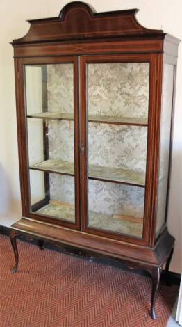 An Edwardian mahogany display cabinet, surmounted by an arched pediment, with boxwood stringing above two glazed doors and fitted shelves, on cabriole legs terminating in club feet, 191cm H, 107cm W, 41cm D.