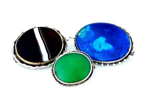 Three silver natural stone set brooches, to include a bull's eye agate brooch, in wavy design silver border, 3cm W, a lapis lazuli oval brooch, in silver reeded design border, 3.2cm W, and a green jade oval brooch, 2.4cm W. (3)