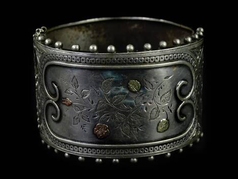 A Victorian aesthetic silver bangle, with tricolour design leaves and flowers on central panel, with beaded border, and safety chain, 6cm W.