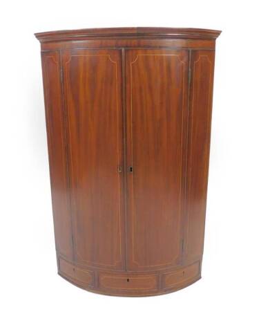 A George III mahogany bowfront hanging corner cupboard, with moulded cornice and three shelves enclosed by two doors with boxwood stringing and fitted with a drawer below, 111cm H, 73cm W.