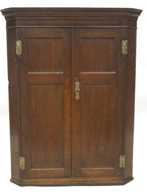 A George III oak hanging corner cupboard, with moulded cornice over two doors, each with moulded panelled doors, H hinges and brass escutcheon revealing shaped shelves, 106cm H, 84cm W across the canted sides.