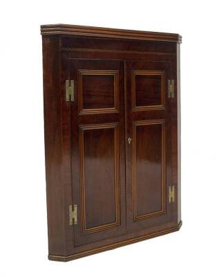 A George III mahogany hanging corner cupboard, with triple moulded cornice, ebony and boxwood banding to the frieze having two double panel doors of conforming decoration, on brass H hinges enclosing shaped shelves, 105cm H, 80cm W across the canted sides