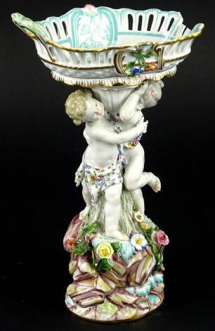 A Meissen porcelain centrepiece, the basket shaped bowl with pierced sides and handles, on a double figural column support, with stylised rock base, embellished with flower encrusted detail, blue cross marks to underside (AF), 26cm H.