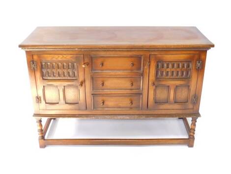 A Reprodux oak sideboard, with a pair of paneled doors flanking three drawers, raised on turned supports and square block feet, united by stretchers, 86cm H, 128cm W, 47cm D.