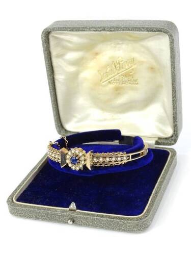 An Edwardian hinged bangle, set with central cluster of sapphire surrounded by seed pearls, with fine ball and wire design, and seed pearl set shoulders, yellow metal, unmarked, with safety chain, 6cm wide.