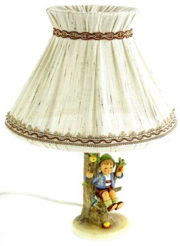 A Goebel Hummel ceramic lamp base, modelled in the form of a boy seated on a fruit tree branch beside a bird, with shade, 44cm H overall.