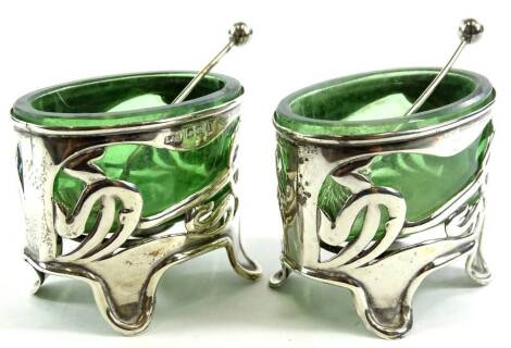 A pair of Edwardian silver salts, of Art Nouveau form, with pierced floral bodies on quadruple feet, with oval green glass liners, and associated spoons, Birmingham 1908.